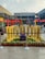 A row of large, yellow cricket bats is displayed outdoors, each with the name of an Indian city printed on it. The setting is a shopping plaza with storefronts like Buffalo Wild Wings and Barley & Grapes Brewery visible in the background. Red umbrellas and small potted plants decorate the area. The space is cordoned off with blue barriers, and a small table or box in the center mentions a live screening at Aero Plaza.