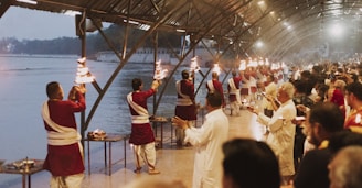 A group of people dressed in traditional attire are performing a ritual along a riverbank under a large covered structure. They are holding lit torches and appear to be in a coordinated sequence. Numerous onlookers are gathered, observing the ritual, contributing to a serene and respectful atmosphere. The background features a calm river and buildings, suggesting a religious or cultural setting.