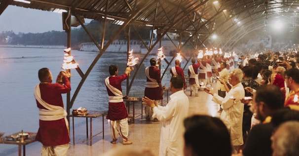 A group of people dressed in traditional attire are performing a ritual along a riverbank under a large covered structure. They are holding lit torches and appear to be in a coordinated sequence. Numerous onlookers are gathered, observing the ritual, contributing to a serene and respectful atmosphere. The background features a calm river and buildings, suggesting a religious or cultural setting.