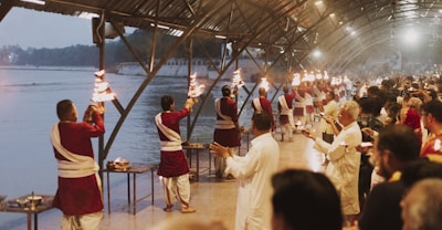 A group of people dressed in traditional attire are performing a ritual along a riverbank under a large covered structure. They are holding lit torches and appear to be in a coordinated sequence. Numerous onlookers are gathered, observing the ritual, contributing to a serene and respectful atmosphere. The background features a calm river and buildings, suggesting a religious or cultural setting.