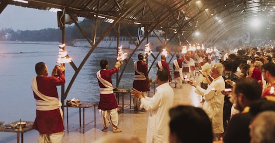 A group of people dressed in traditional attire are performing a ritual along a riverbank under a large covered structure. They are holding lit torches and appear to be in a coordinated sequence. Numerous onlookers are gathered, observing the ritual, contributing to a serene and respectful atmosphere. The background features a calm river and buildings, suggesting a religious or cultural setting.