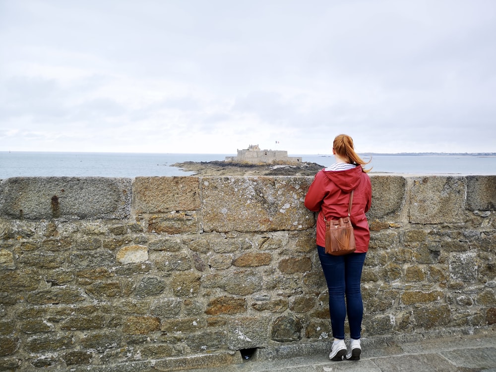 a woman leaning against a stone wall near the ocean