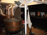 A room with industrial-style metal vats, some open with wooden paddles inside, surrounded by white pipes and other equipment. There are copper pots on a black stove or counter in the background, and a fabric item hanging nearby.