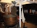 A room with industrial-style metal vats, some open with wooden paddles inside, surrounded by white pipes and other equipment. There are copper pots on a black stove or counter in the background, and a fabric item hanging nearby.