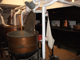 A room with industrial-style metal vats, some open with wooden paddles inside, surrounded by white pipes and other equipment. There are copper pots on a black stove or counter in the background, and a fabric item hanging nearby.