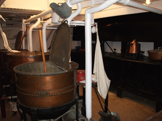 A room with industrial-style metal vats, some open with wooden paddles inside, surrounded by white pipes and other equipment. There are copper pots on a black stove or counter in the background, and a fabric item hanging nearby.