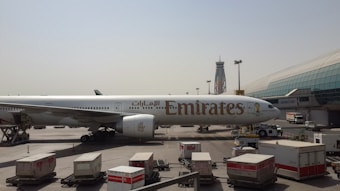 An Emirates airplane is parked on the tarmac at an airport, with the terminal building visible in the background. Several cargo containers and ground support vehicles surround the aircraft. A tall control tower with colorful design elements can be seen in the distance under a clear sky.