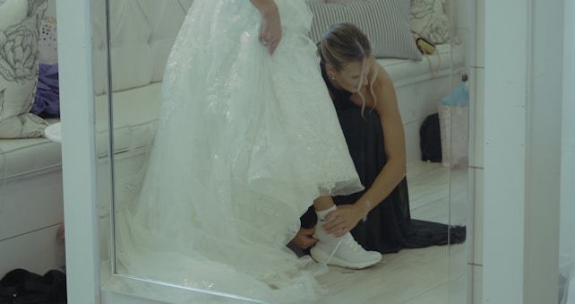 A woman dressed in a black outfit is helping another person, likely a bride, put on white sneakers under a wedding dress. They are in a room with white and beige decor, including a sofa with floral-patterned cushions.