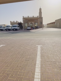 A small building with a tall tower-like structure on the right, possibly a mosque or community center, is surrounded by several parked vans and a car. The building has a sandy beige color, and there is a wide tiled courtyard leading up to it. The environment appears quiet and sunlit, suggesting a calm and serene setting.