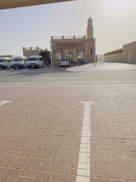 A small building with a tall tower-like structure on the right, possibly a mosque or community center, is surrounded by several parked vans and a car. The building has a sandy beige color, and there is a wide tiled courtyard leading up to it. The environment appears quiet and sunlit, suggesting a calm and serene setting.