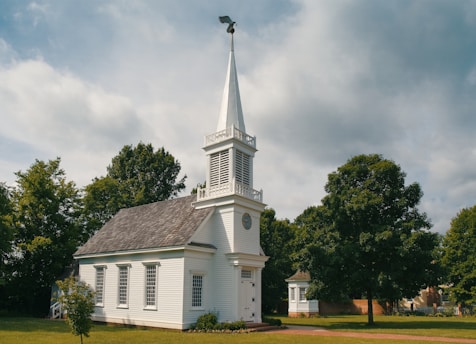 A small white church with a tall steeple topped by a weather vane, surrounded by lush green trees. The building has several windows and is set on a well-kept lawn under a partly cloudy sky.