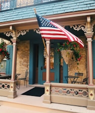 A quaint and charming front porch of a building featuring intricate woodwork and a hanging American flag. The space has a cozy atmosphere with wicker baskets filled with vibrant red flowers and foliage. A pair of simple black metal chairs and tables are placed on the porch, complementing the warm-toned brick wall and the teal-colored accents of the doors and shutters.