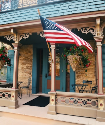 A quaint and charming front porch of a building featuring intricate woodwork and a hanging American flag. The space has a cozy atmosphere with wicker baskets filled with vibrant red flowers and foliage. A pair of simple black metal chairs and tables are placed on the porch, complementing the warm-toned brick wall and the teal-colored accents of the doors and shutters.