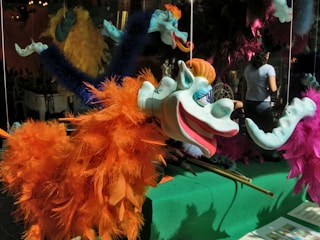 A vibrant puppet with a whimsical design features a blue face, large eyes, and a wide smile. It is adorned with bright orange and blue feathers, hanging by strings. In the background, there are other colorful puppets, and a person stands nearby, slightly obscured.