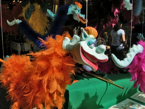 A vibrant puppet with a whimsical design features a blue face, large eyes, and a wide smile. It is adorned with bright orange and blue feathers, hanging by strings. In the background, there are other colorful puppets, and a person stands nearby, slightly obscured.