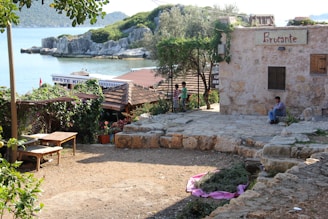 A quaint seaside scene featuring a rustic stone building with a sign reading 'Brocante.' A man sits on a step nearby, while two children stand near a fence. The area is surrounded by greenery, including vines, trees, and potted plants. The background reveals a calm sea with rocky outcrops and distant hills. A partially visible sign reads 'Beste Kume,' suggesting the presence of a nearby establishment or market.