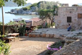 A quaint seaside scene featuring a rustic stone building with a sign reading 'Brocante.' A man sits on a step nearby, while two children stand near a fence. The area is surrounded by greenery, including vines, trees, and potted plants. The background reveals a calm sea with rocky outcrops and distant hills. A partially visible sign reads 'Beste Kume,' suggesting the presence of a nearby establishment or market.