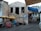 A rustic stone building with two large window openings. Below it, there are colorful market stalls with yellow and red umbrellas displaying textiles and trinkets. A sign reads 'Kabay Open Air Bar' and 'Free Internet'. Nearby, a person sits next to the stall under an umbrella. There are several buildings in the background, one with a blue canopy.
