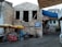A rustic stone building with two large window openings. Below it, there are colorful market stalls with yellow and red umbrellas displaying textiles and trinkets. A sign reads 'Kabay Open Air Bar' and 'Free Internet'. Nearby, a person sits next to the stall under an umbrella. There are several buildings in the background, one with a blue canopy.