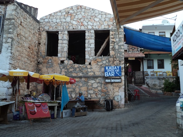 A rustic stone building with two large window openings. Below it, there are colorful market stalls with yellow and red umbrellas displaying textiles and trinkets. A sign reads 'Kabay Open Air Bar' and 'Free Internet'. Nearby, a person sits next to the stall under an umbrella. There are several buildings in the background, one with a blue canopy.