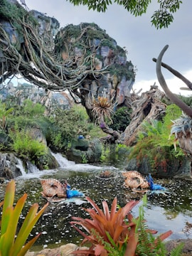 A lush, fantastical landscape with intricate, floating rock formations covered in dense foliage and vines. Below, there is a serene waterway with small waterfalls and vibrant, unusual plants, including striking blue and orange foliage. The scene evokes a sense of otherworldly beauty and tranquil mystery.