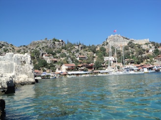 A scenic coastal village nestled in a rocky hillside, featuring numerous small houses with red-tiled roofs and surrounded by lush greenery. A prominent stone fortress with a flag is perched atop the hill, overlooking the calm, turquoise sea. Several sailboats are docked along the shoreline, enhancing the picturesque landscape.