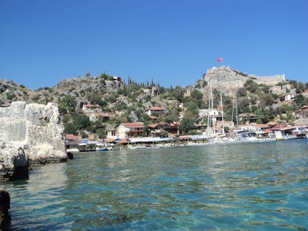 A scenic coastal village nestled in a rocky hillside, featuring numerous small houses with red-tiled roofs and surrounded by lush greenery. A prominent stone fortress with a flag is perched atop the hill, overlooking the calm, turquoise sea. Several sailboats are docked along the shoreline, enhancing the picturesque landscape.