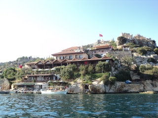 A picturesque view of a hillside settlement by the water, featuring buildings with red-tiled roofs nestled among rocky terrain and greenery. Several Turkish flags are visible, indicating a Turkish location. A few boats are docked along the shoreline, adding to the serene coastal atmosphere.