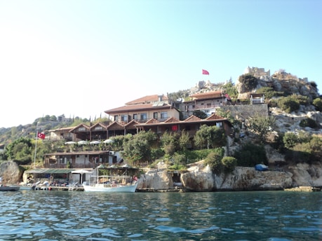 A picturesque view of a hillside settlement by the water, featuring buildings with red-tiled roofs nestled among rocky terrain and greenery. Several Turkish flags are visible, indicating a Turkish location. A few boats are docked along the shoreline, adding to the serene coastal atmosphere.