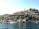 A picturesque view of a hillside settlement by the water, featuring buildings with red-tiled roofs nestled among rocky terrain and greenery. Several Turkish flags are visible, indicating a Turkish location. A few boats are docked along the shoreline, adding to the serene coastal atmosphere.