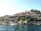 A picturesque view of a hillside settlement by the water, featuring buildings with red-tiled roofs nestled among rocky terrain and greenery. Several Turkish flags are visible, indicating a Turkish location. A few boats are docked along the shoreline, adding to the serene coastal atmosphere.