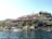 A picturesque view of a hillside settlement by the water, featuring buildings with red-tiled roofs nestled among rocky terrain and greenery. Several Turkish flags are visible, indicating a Turkish location. A few boats are docked along the shoreline, adding to the serene coastal atmosphere.