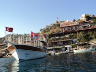 A wooden boat with a Turkish flag is docked by a scenic hillside. The hillside features a series of quaint cafes and stone buildings surrounded by greenery. The sky is clear, providing a serene backdrop, and there are additional boats in the water.