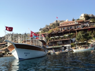 A wooden boat with a Turkish flag is docked by a scenic hillside. The hillside features a series of quaint cafes and stone buildings surrounded by greenery. The sky is clear, providing a serene backdrop, and there are additional boats in the water.