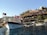 A wooden boat with a Turkish flag is docked by a scenic hillside. The hillside features a series of quaint cafes and stone buildings surrounded by greenery. The sky is clear, providing a serene backdrop, and there are additional boats in the water.