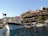 A wooden boat with a Turkish flag is docked by a scenic hillside. The hillside features a series of quaint cafes and stone buildings surrounded by greenery. The sky is clear, providing a serene backdrop, and there are additional boats in the water.