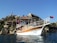 A wooden boat with a white hull and Turkish flags docked near a rocky shore. Behind the boat, there are houses and a small hill with a few trees. A larger fortress or castle is visible at the top of the hill.