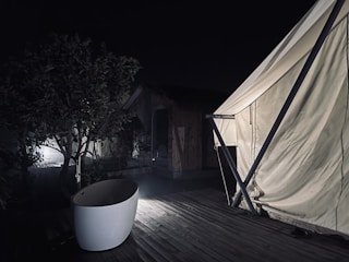 A nighttime scene featuring a partially lit outdoor bathtub on a wooden deck next to a large white tent. A tree and wooden cabin are visible in the dim background.