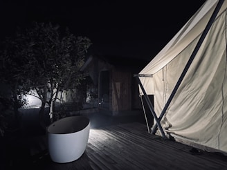 A nighttime scene featuring a partially lit outdoor bathtub on a wooden deck next to a large white tent. A tree and wooden cabin are visible in the dim background.