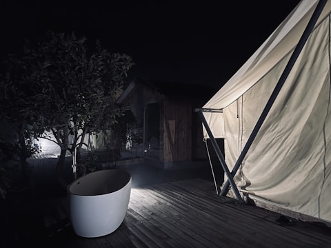 A nighttime scene featuring a partially lit outdoor bathtub on a wooden deck next to a large white tent. A tree and wooden cabin are visible in the dim background.