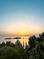 A serene Greek island sunset over the sea with lush greenery in the foreground.