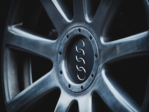 A close-up view of a car wheel with a metallic alloy finish, featuring a central logo of four interlinked rings. The rim has a sleek, modern design with visible bolt patterns around the center.