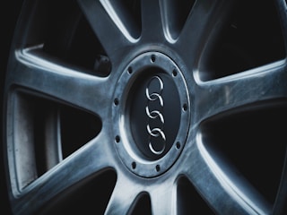 A close-up view of a car wheel with a metallic alloy finish, featuring a central logo of four interlinked rings. The rim has a sleek, modern design with visible bolt patterns around the center.