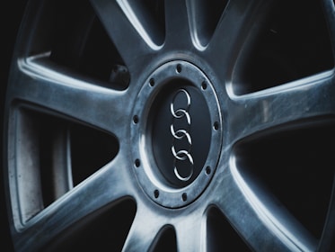 A close-up view of a car wheel with a metallic alloy finish, featuring a central logo of four interlinked rings. The rim has a sleek, modern design with visible bolt patterns around the center.