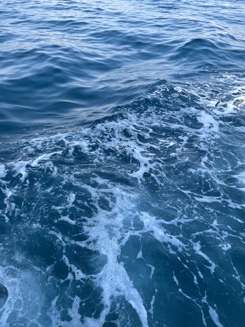 Waves and ripples on a body of water with visible foam and bubbles. The water appears deep blue with lighter areas where the foam is present. The surface is slightly rough, indicating mild turbulence.