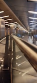 A modern architectural interior featuring a sleek, metallic railing that leads down a long corridor with a series of lights along the ceiling. The space is well-lit with reflective surfaces, and the design emphasizes clean lines and symmetry, creating a futuristic atmosphere.
