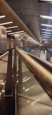A modern architectural interior featuring a sleek, metallic railing that leads down a long corridor with a series of lights along the ceiling. The space is well-lit with reflective surfaces, and the design emphasizes clean lines and symmetry, creating a futuristic atmosphere.