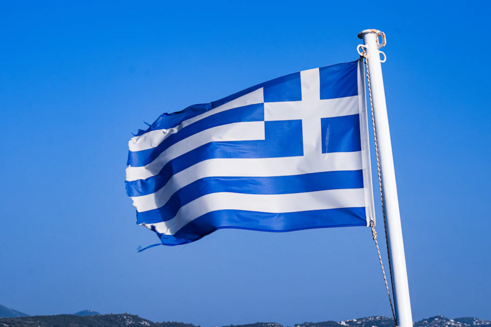Greek flag flying high on white pole against clear blue sky with waving motion
