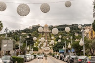 A landscape view of a well-designed garden with terraced levels and a prominent, golden-domed building at the top. The garden features a series of large, decorative, white spherical ornaments hanging overhead. Trees are symmetrically planted along the terraced slopes, creating an organized and lush appearance. Cars are parked along the road leading to the entrance of the garden.