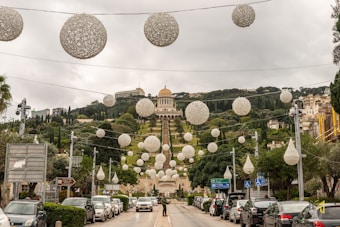 A landscape view of a well-designed garden with terraced levels and a prominent, golden-domed building at the top. The garden features a series of large, decorative, white spherical ornaments hanging overhead. Trees are symmetrically planted along the terraced slopes, creating an organized and lush appearance. Cars are parked along the road leading to the entrance of the garden.